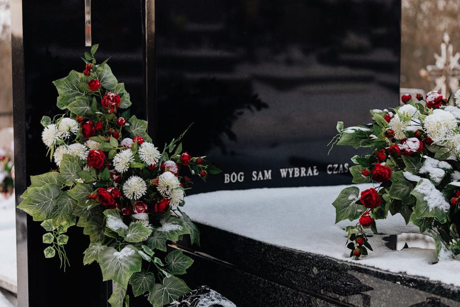 Elegant funeral tribute featuring a symmetrical arrangement of fresh red roses, white chrysanthemums, and green holly leaves, with some petals and leaves lightly dusted with snow. The flowers are plac
